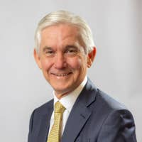 Smiling elderly man in a dark suit and patterned tie, head-and-shoulders portrait against a plain gray background