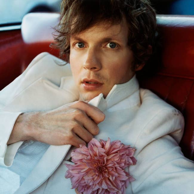Person in a white suit with a pink flower in the lapel, sitting on a red seat, looking directly at the camera.