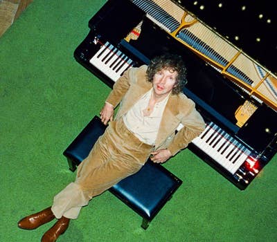 Person in a tan suit sits on a piano bench, viewed from above, with a grand piano and green carpet in the background.