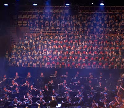 Large choir in matching shirts performs above an orchestra on stage, with a conductor leading the musicians below.