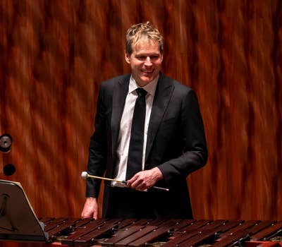 Man in a suit playing a marimba with a mallet, standing on a stage with a wooden backdrop.