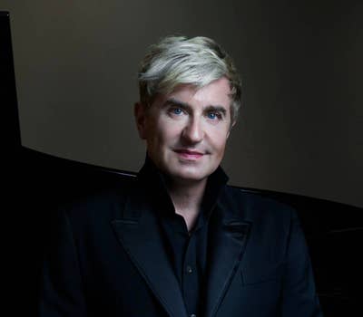 Man with short blond hair wearing a black suit, standing in front of a dark background.
