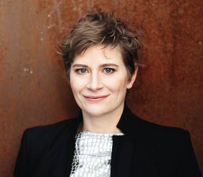 Smiling woman with short hair wearing a black jacket and patterned top, standing against a rust-colored background.