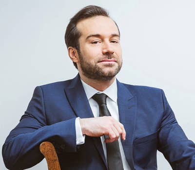 Man in a blue suit and black tie sitting on a wooden chair, looking confidently at the camera against a plain background.