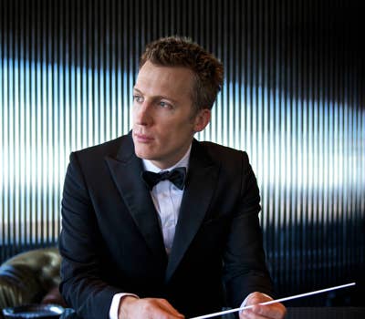 Man in a tuxedo holding a conductor's baton, seated at a table with a striped background.