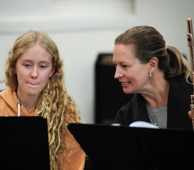 Woman with flute instructs a young girl in a music lesson, both focused on sheet music in a classroom setting.