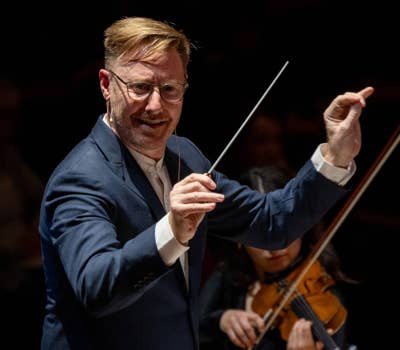 Conductor in a blue suit leading an orchestra, with a violinist in the background.