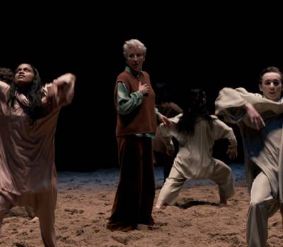 Dancers perform expressively on a sandy stage, wearing loose, neutral-toned clothing, with a dark background.