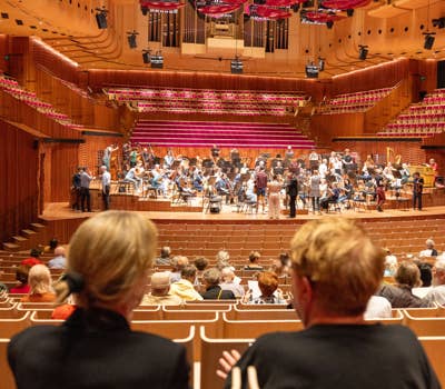 Orchestra rehearsing on stage in a concert hall with an audience seated in the foreground.