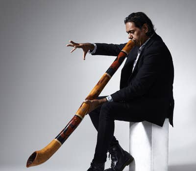Man in a black suit playing a decorated didgeridoo while seated on a white block against a gray background.